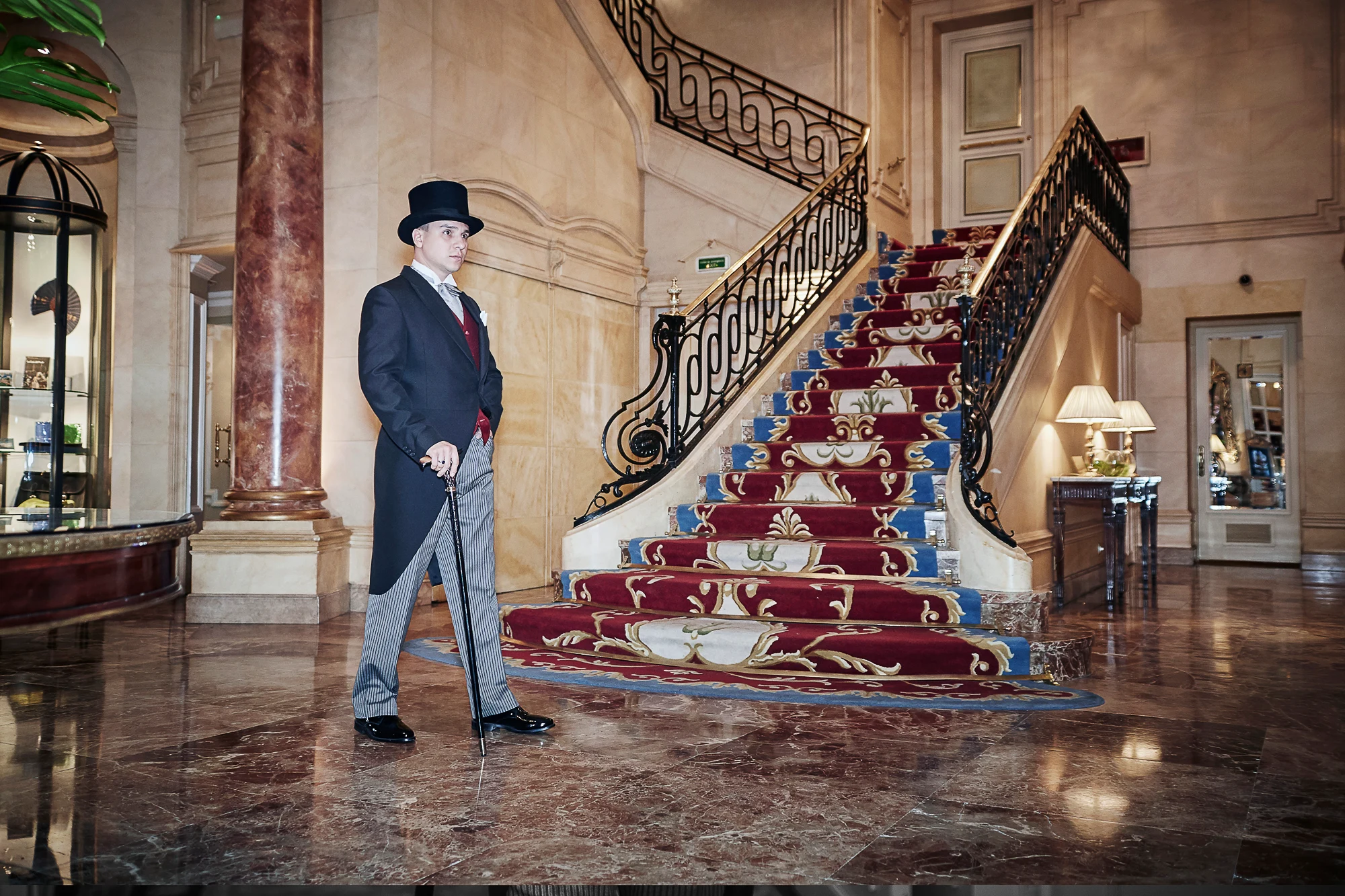 Gentleman in a top hat and tailcoat standing in an ornate hotel lobby with marble staircase (Pavel Manylov)