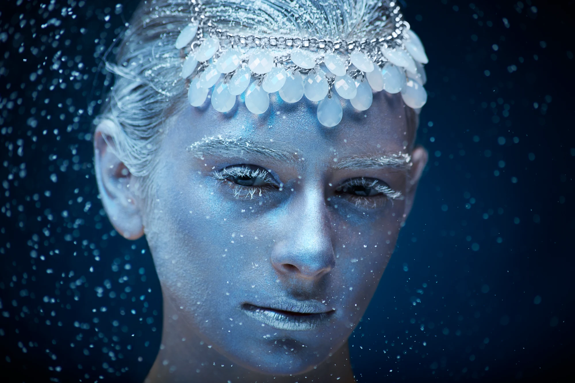 Close up fantasy portrait with blue icy makeup, white brows and headpiece in falling snow