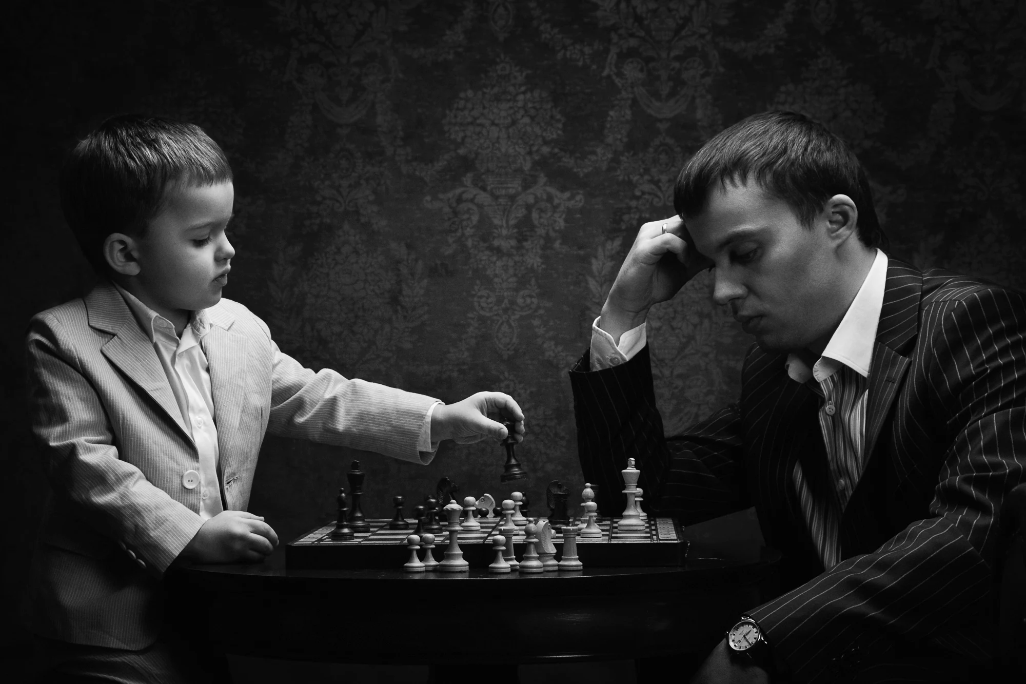 Little boy in suit playing chess with his father in dark vintage studio