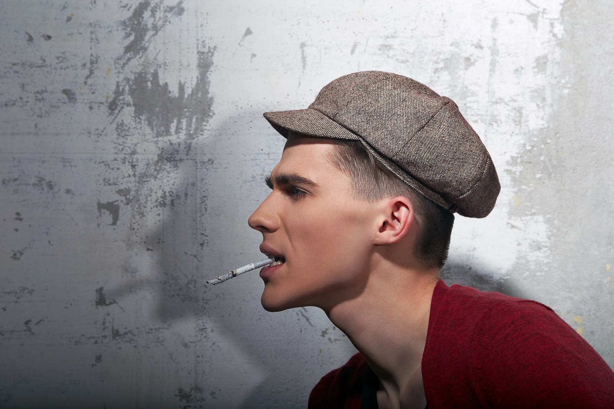 Young man in flat cap smoking a cigarette in profile against concrete wall
