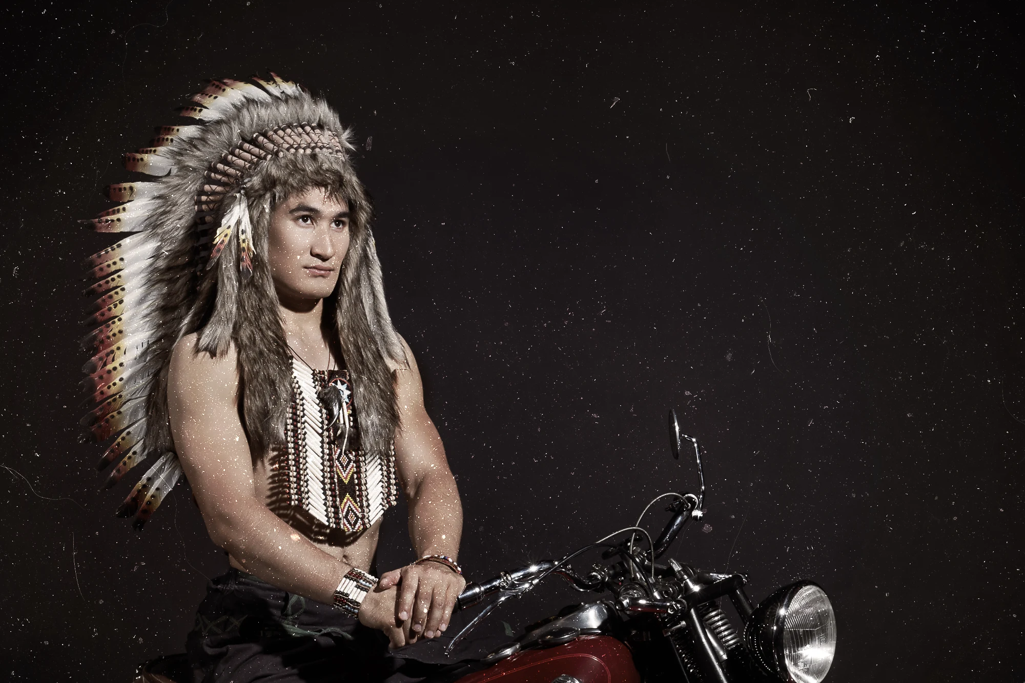 Native American chief wearing feather headdress sitting on an Indian motorcycle in dark studio setting