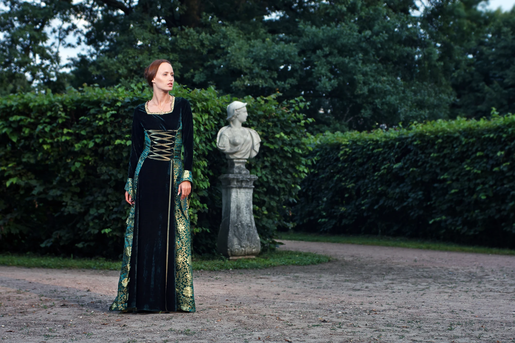 Woman in long medieval green velvet dress standing in classical garden with statue