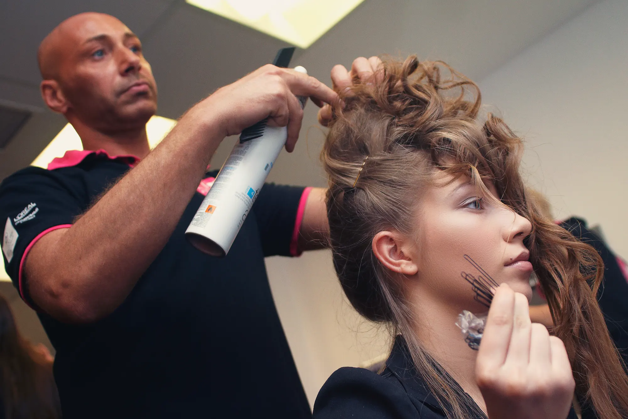 Hairstylist spraying and shaping an intricate updo for a young woman backstage before an event in Italy