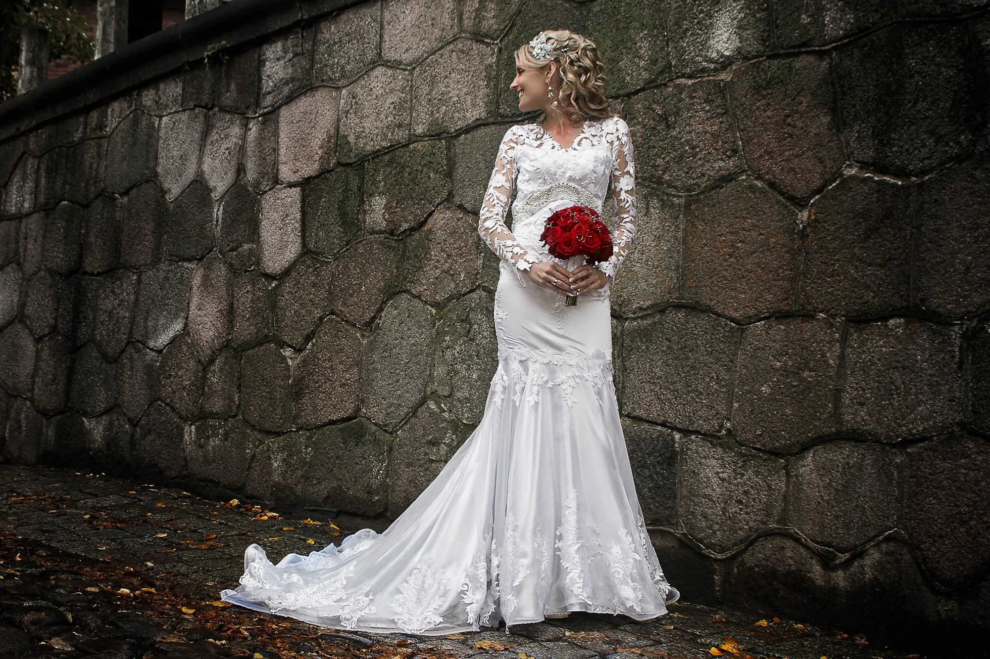 Wedding photographer in Europe creating bridal portrait with lace dress and red rose bouquet by stone wall