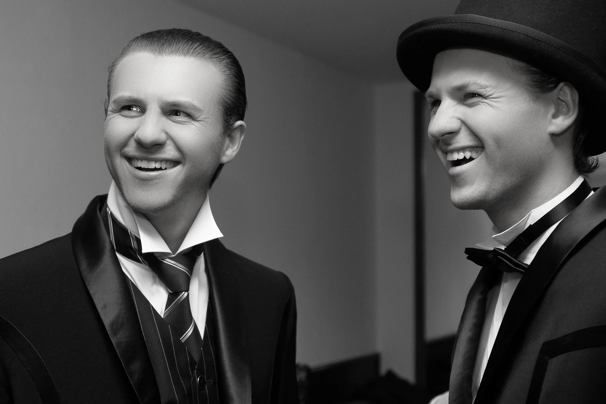 Two men in classic tuxedos laughing and talking together before a formal wedding celebration, black and white portrait