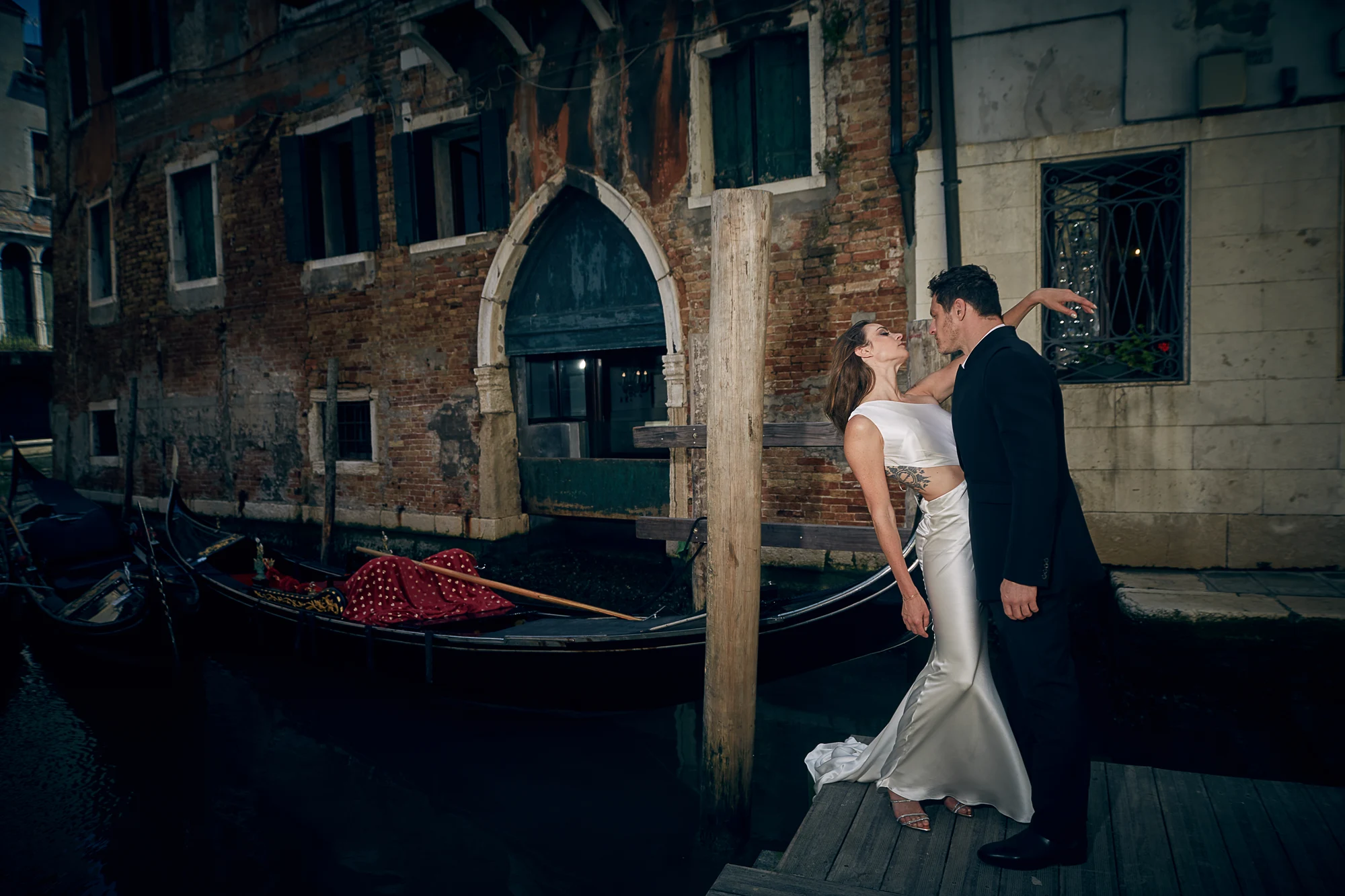 Elegant couple in white dress and black suit posing by gondolas, wedding photographer in Venice Italy