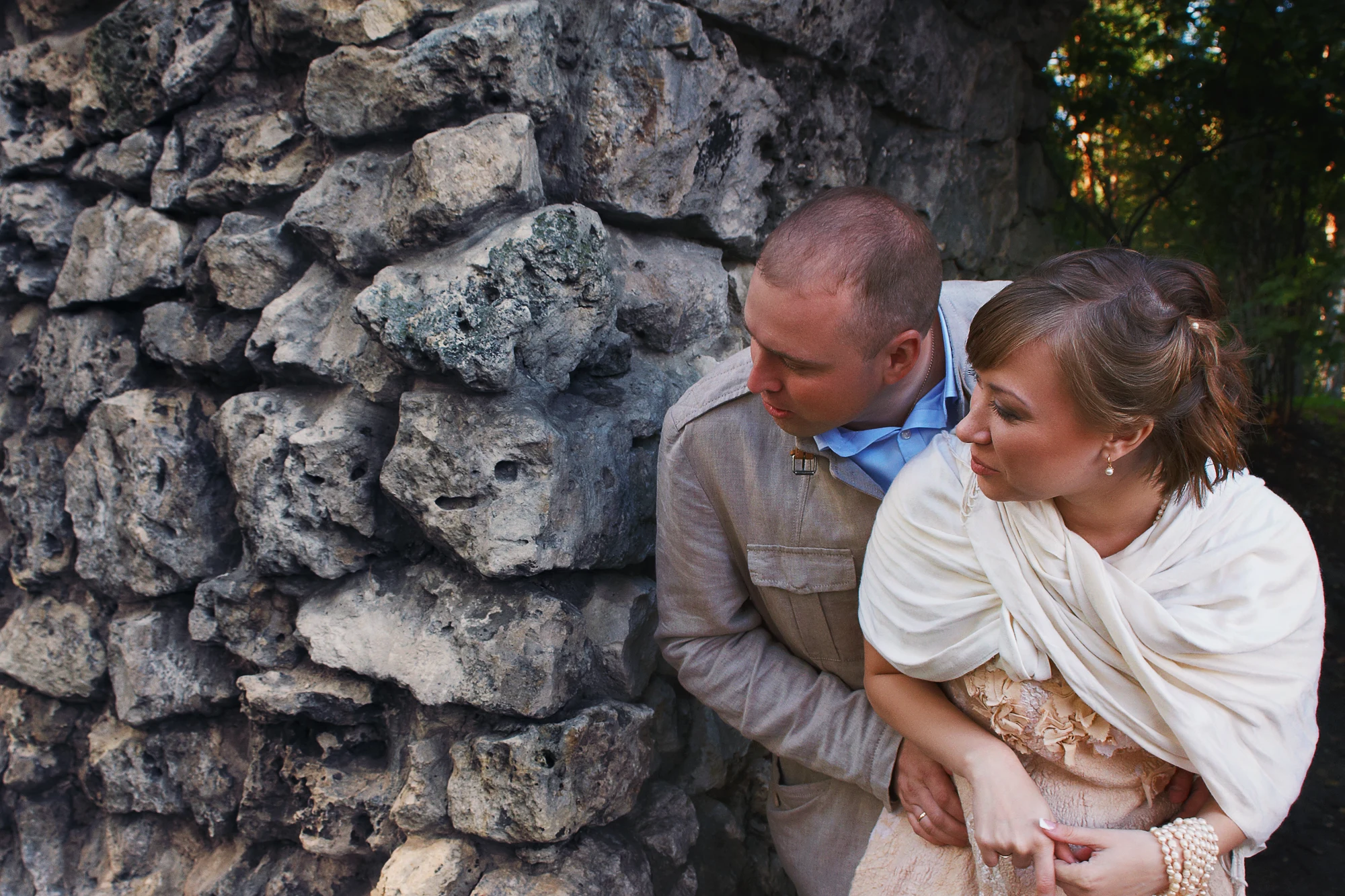 Groom embracing bride wrapped in a light scarf while they stand by an old stone wall in the countryside