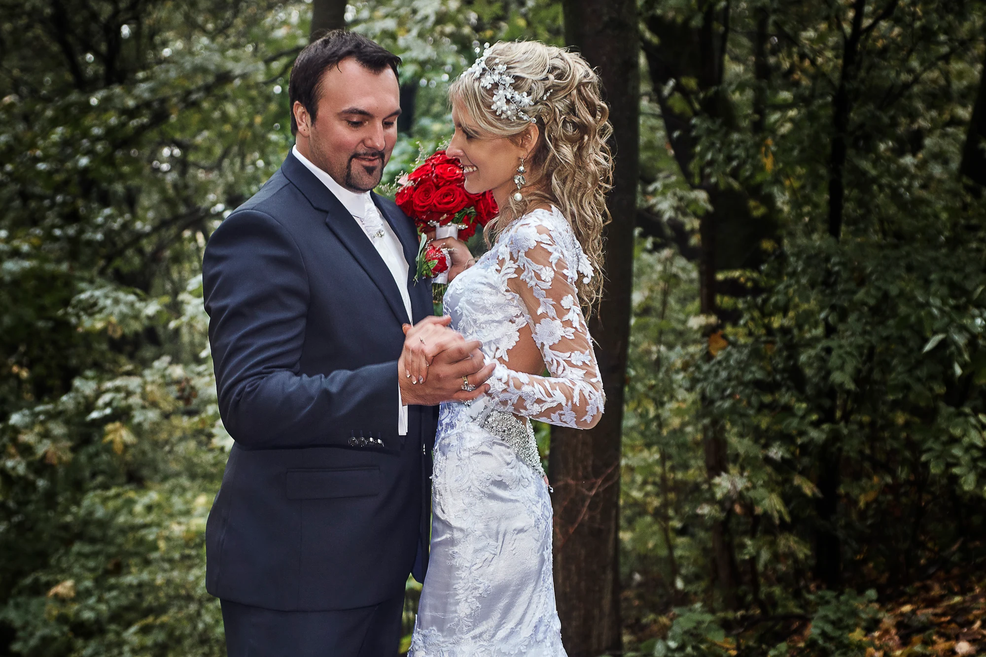 Bride and groom dancing together in a green forest park after rain, the bride holding a bouquet of red roses