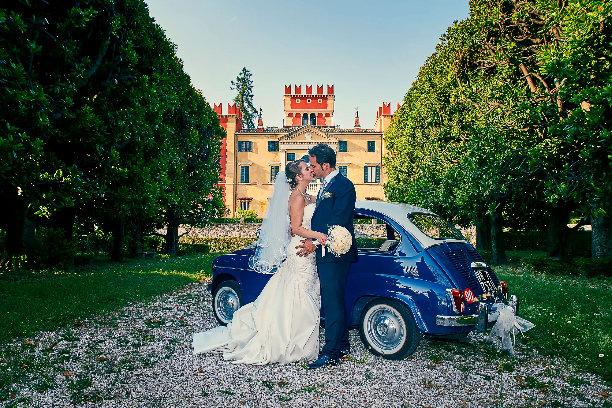 Wedding photographer in Italy shooting bride and groom by vintage blue car in historic villa garden