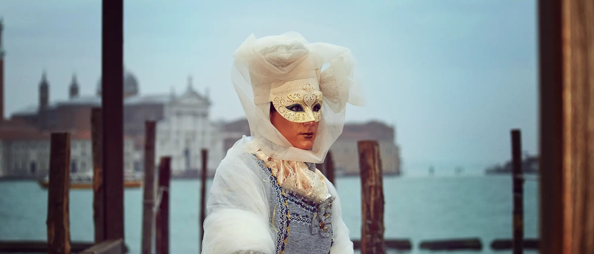 Woman in white Venetian carnival costume and gold mask on pier at sunrise, photographer in Venice